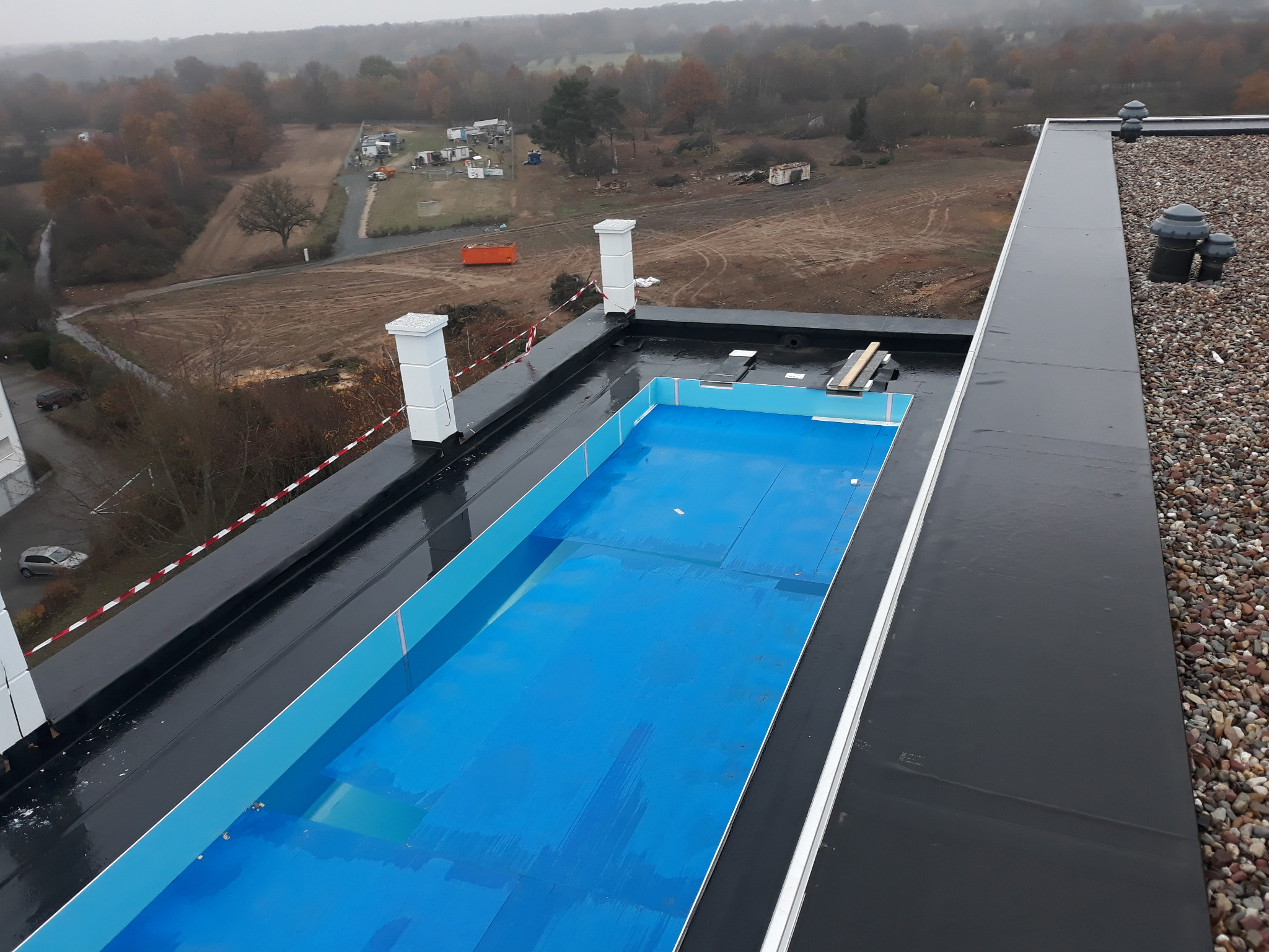 Wykończony dach płaski z niebieską membraną hydroizolacyjną, widoczne kominy wentylacyjne i obróbki blacharskie, w tle jesienny krajobraz.