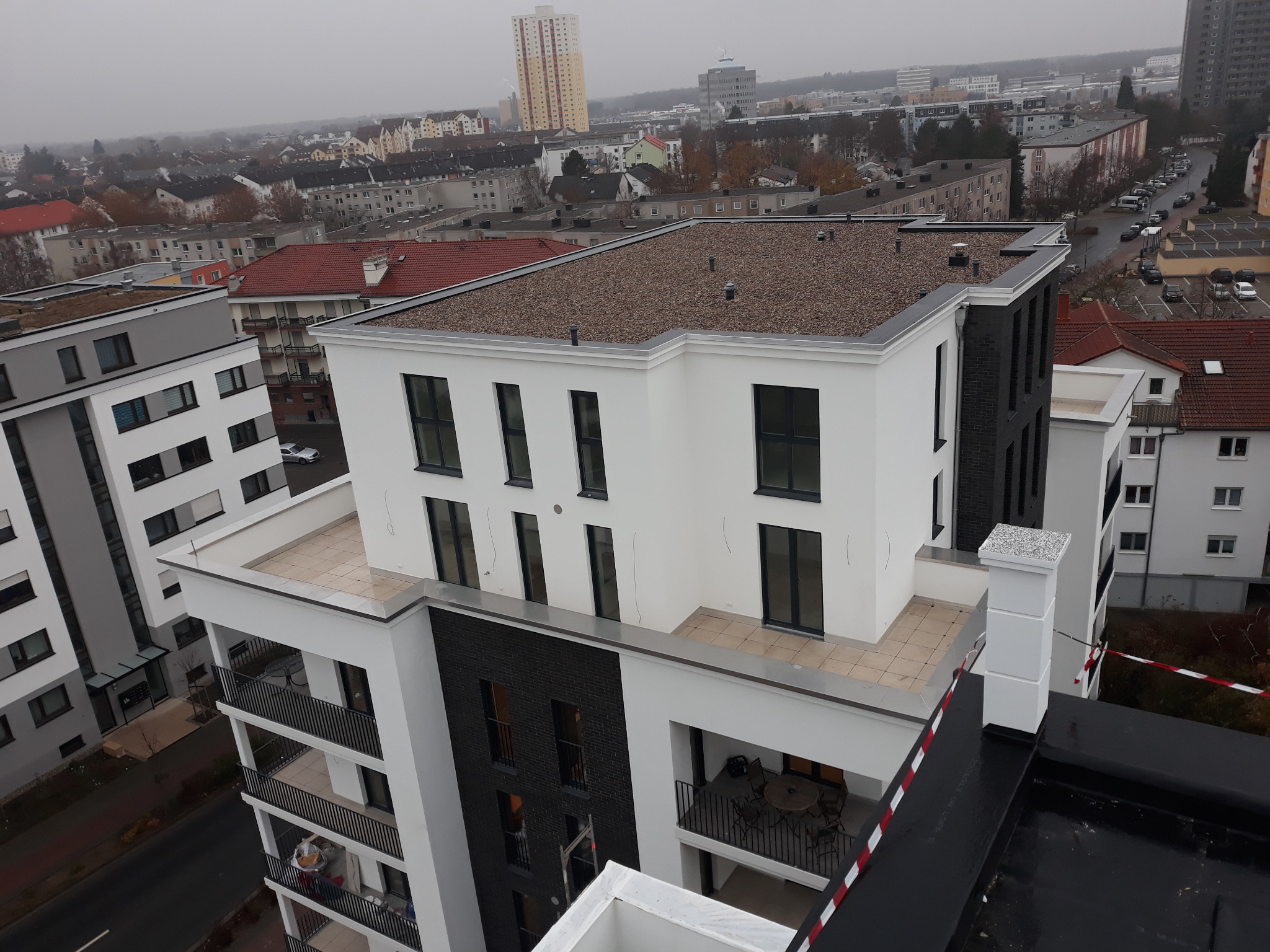 Nowoczesny budynek wielorodzinny z płaskim dachem pokrytym drobnym żwirem, widok z góry na tle miejskiego krajobrazu w pochmurny dzień.