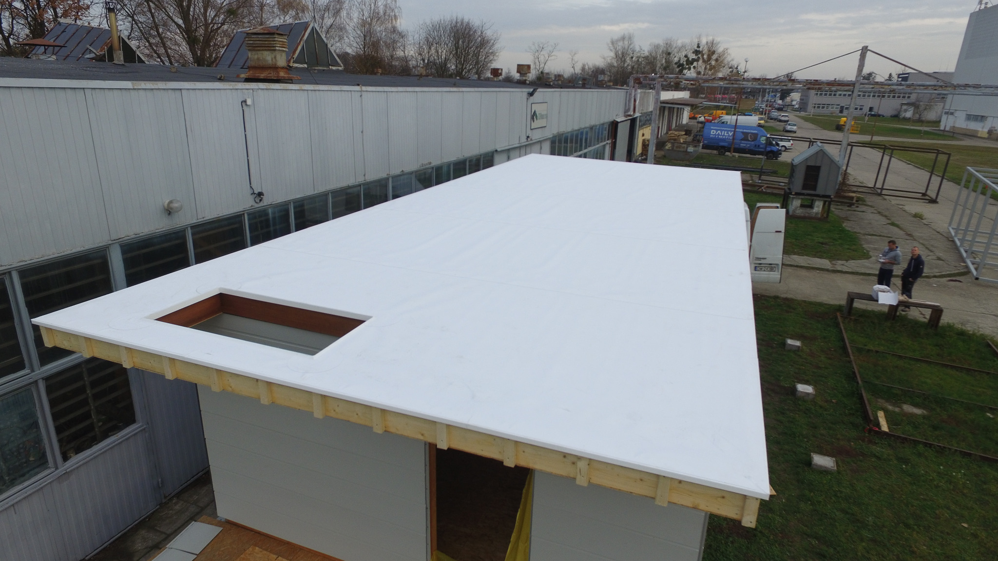 Białe krycie dachu z otworem na świetlik, widoczne drewniane elementy konstrukcyjne, na tle budynków przemysłowych i dwóch osób rozmawiających na zewnątrz.