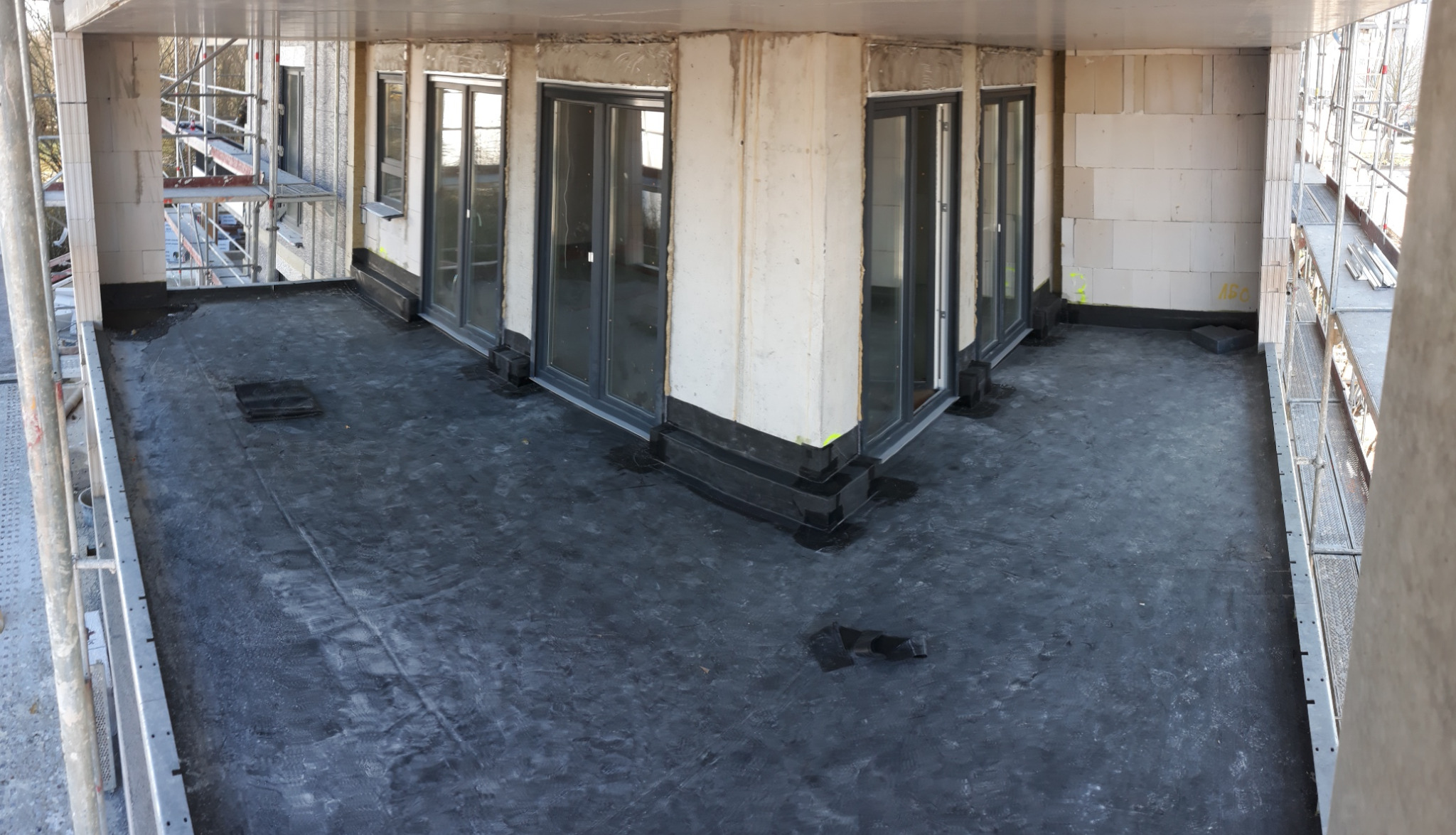 Widok z góry na pokrycie papą termozgrzewalną płaskiego dachu budynku w stanie surowym otwartym, z widocznymi oknami balkonowymi i fragmentami rusztowań.