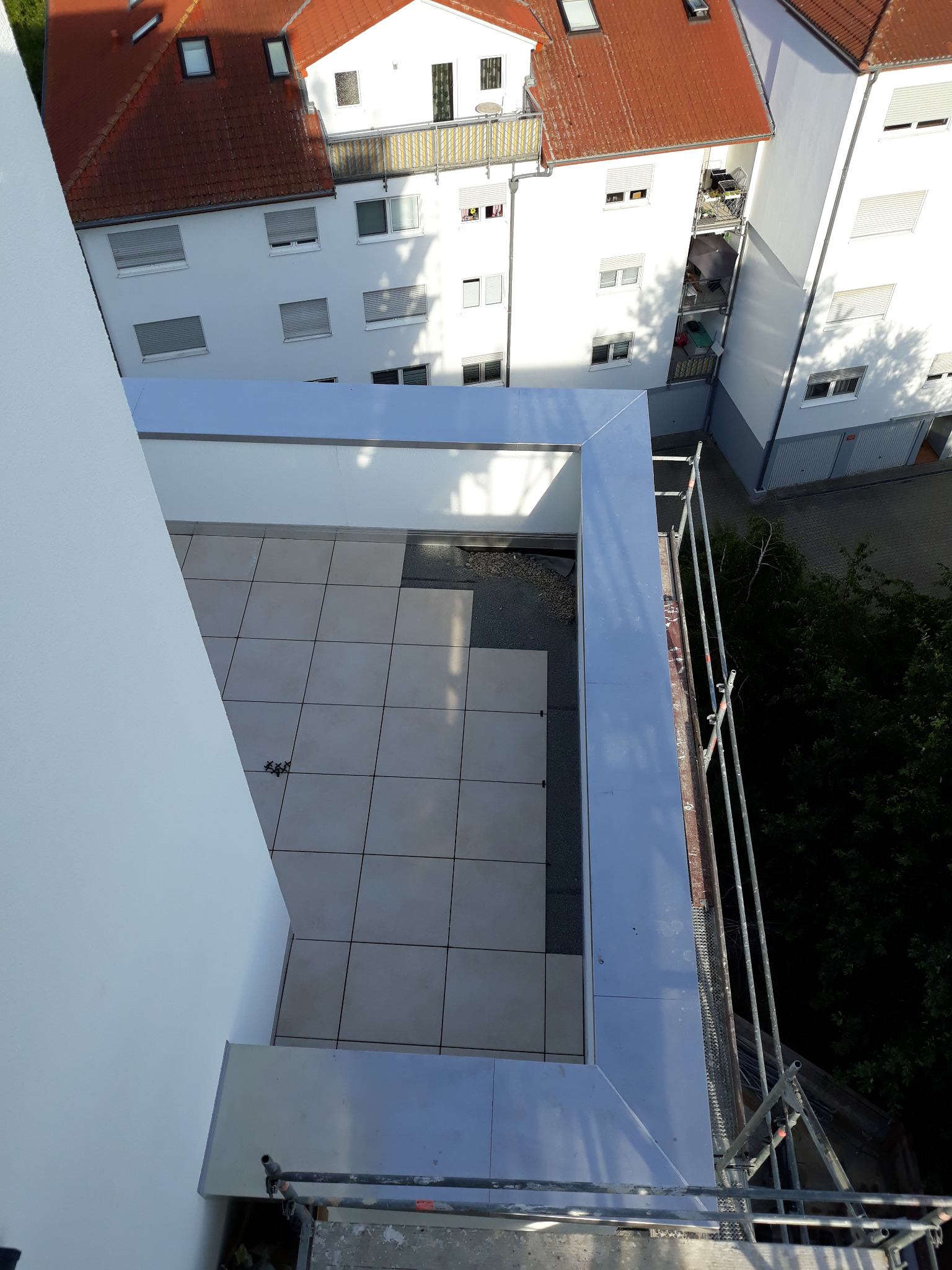 Widok z góry na wykończony dach płaski z szarą ramą i ułożoną szarą płytką, widoczne rusztowanie i budynki w tle.