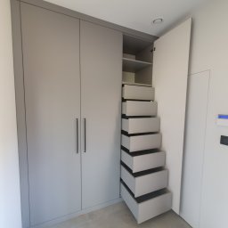 Daniel Bechcicki - Szara szafa wnękowa z otwartymi drzwiami i szeregiem wysuwanych szuflad, widok z przodu na jasnym tle ściany i podłogi.