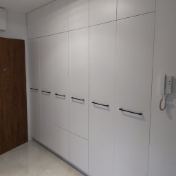 Daniel Bechcicki - Biała, minimalistyczna szafa wnękowa z czarnymi, podłużnymi uchwytami, widoczna w przedpokoju z brązowymi drzwiami i białym domofonem na ścianie.