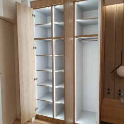 Daniel Bechcicki - Otwarta, trzydrzwiowa szafa wnękowa z oświetleniem LED, jasne drewno i białe wnętrze, widoczne półki i drążek na ubrania, minimalistyczny design, na podłodze panele.