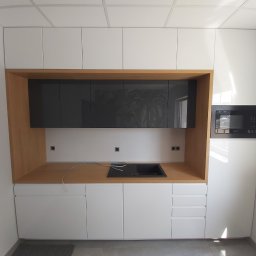 Daniel Bechcicki - Nowoczesna zabudowa kuchenna z białymi szafkami górnymi i dolnymi, drewnianą wnęką z szarymi szafkami i czarnym zlewem, oraz mikrofalówką po prawej stronie, widoczne kable i gniazdka.