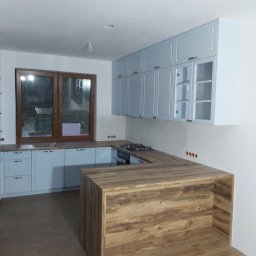 Daniel Bechcicki - Jasna kuchnia z drewnianym blatem wyspowym i błękitnymi szafkami pod oknem, widoczne gniazdka elektryczne na ścianie.