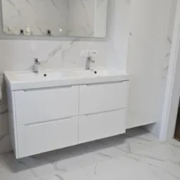 Nowoczesna łazienka z białymi płytkami imitującymi marmur, białą szafką pod umywalką z dwiema umywalkami i bateriami oraz dużym lustrem z oświetleniem LED. Obok szafki znajduje się wysoka, biała...