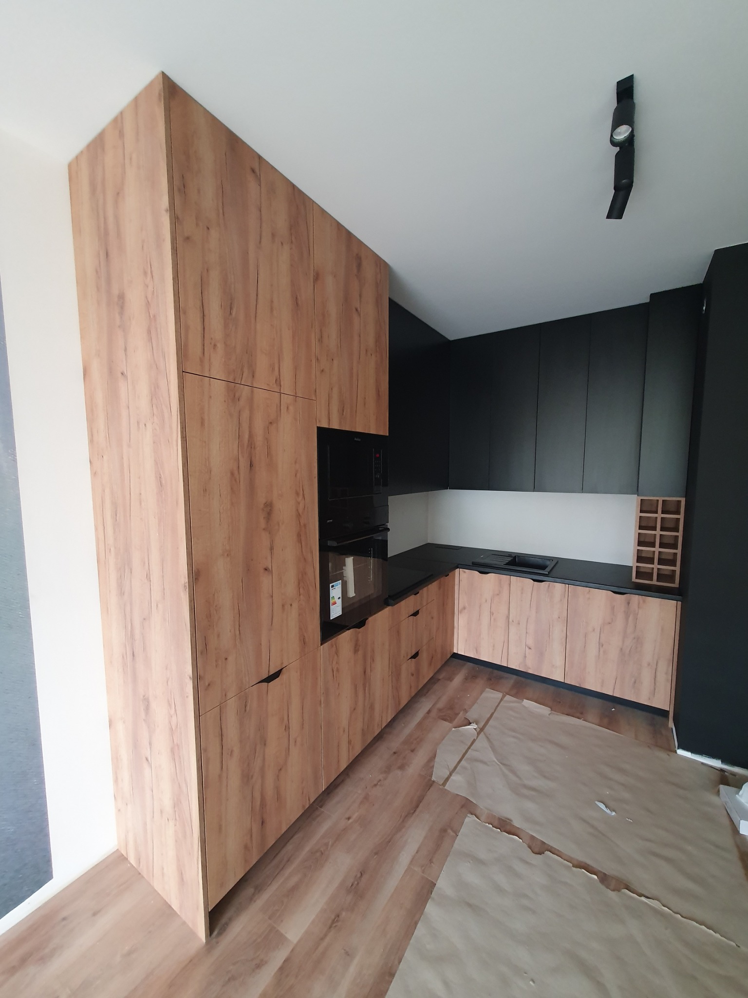 Nowoczesna kuchnia w zabudowie z frontami w odcieniach drewna i czarnymi blatami, widoczne zabezpieczenia podłogi, oświetlenie punktowe sufitowe.