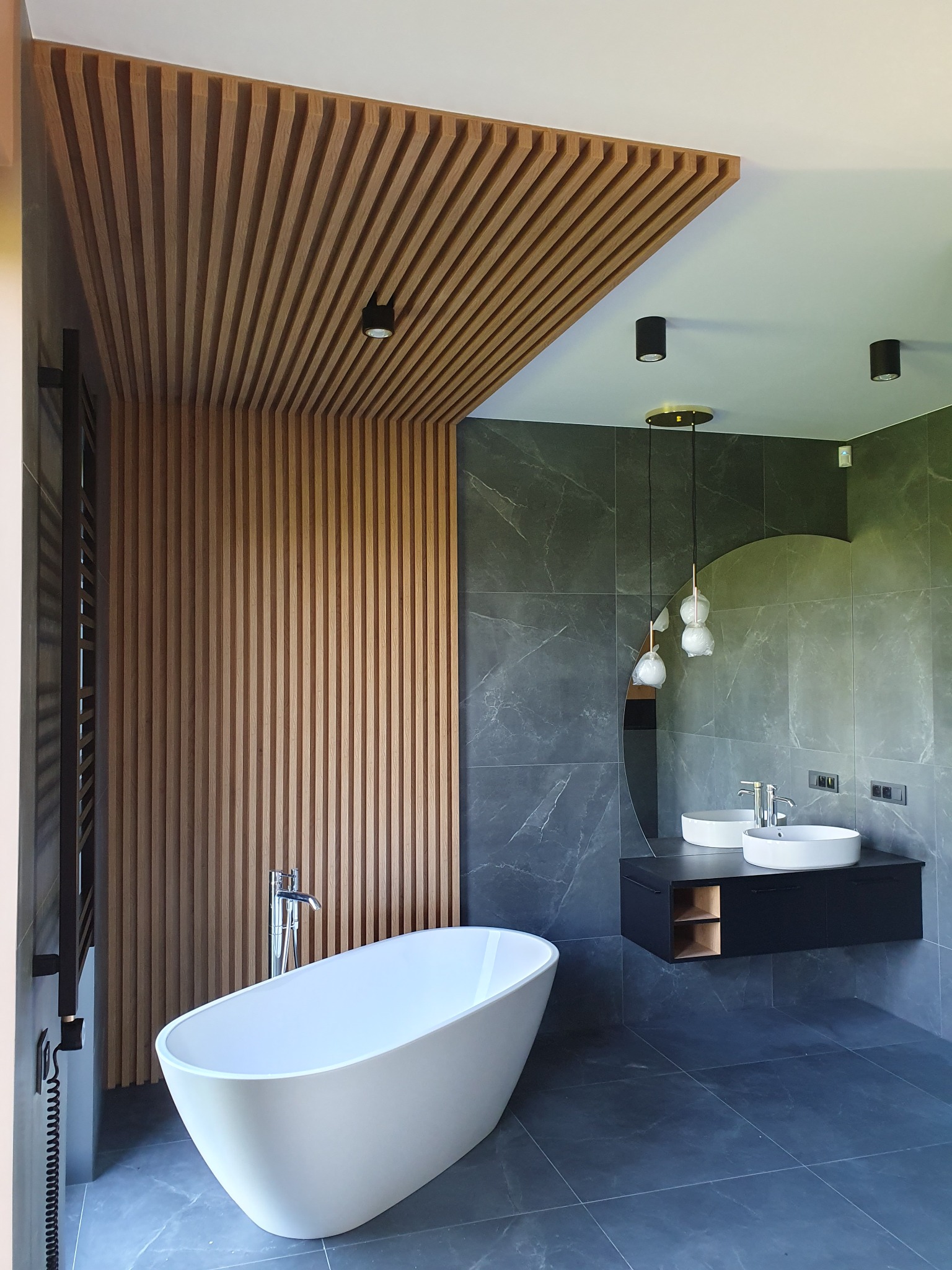 Nowoczesna łazienka z drewnianą okładziną sufitową i ścienną, wolnostojąca wanna, umywalki nablatowe na czarnej szafce, szare płytki imitujące marmur, oświetlenie punktowe i wiszące lampy.