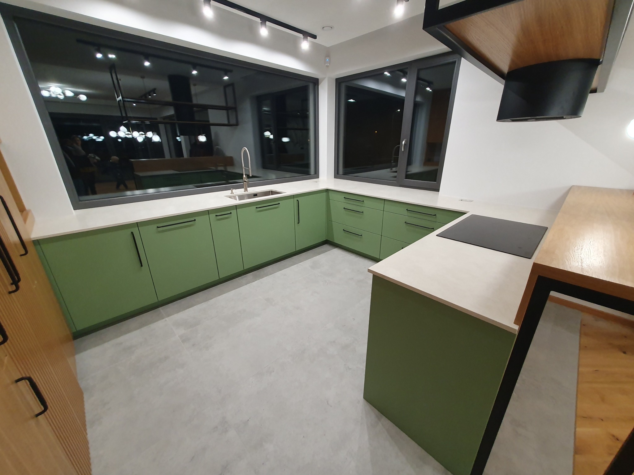 Nowoczesna kuchnia z zielonymi frontami szafek, jasnym blatem i dużymi oknami, widok na oświetlenie sufitowe i fragment drewnianej zabudowy.