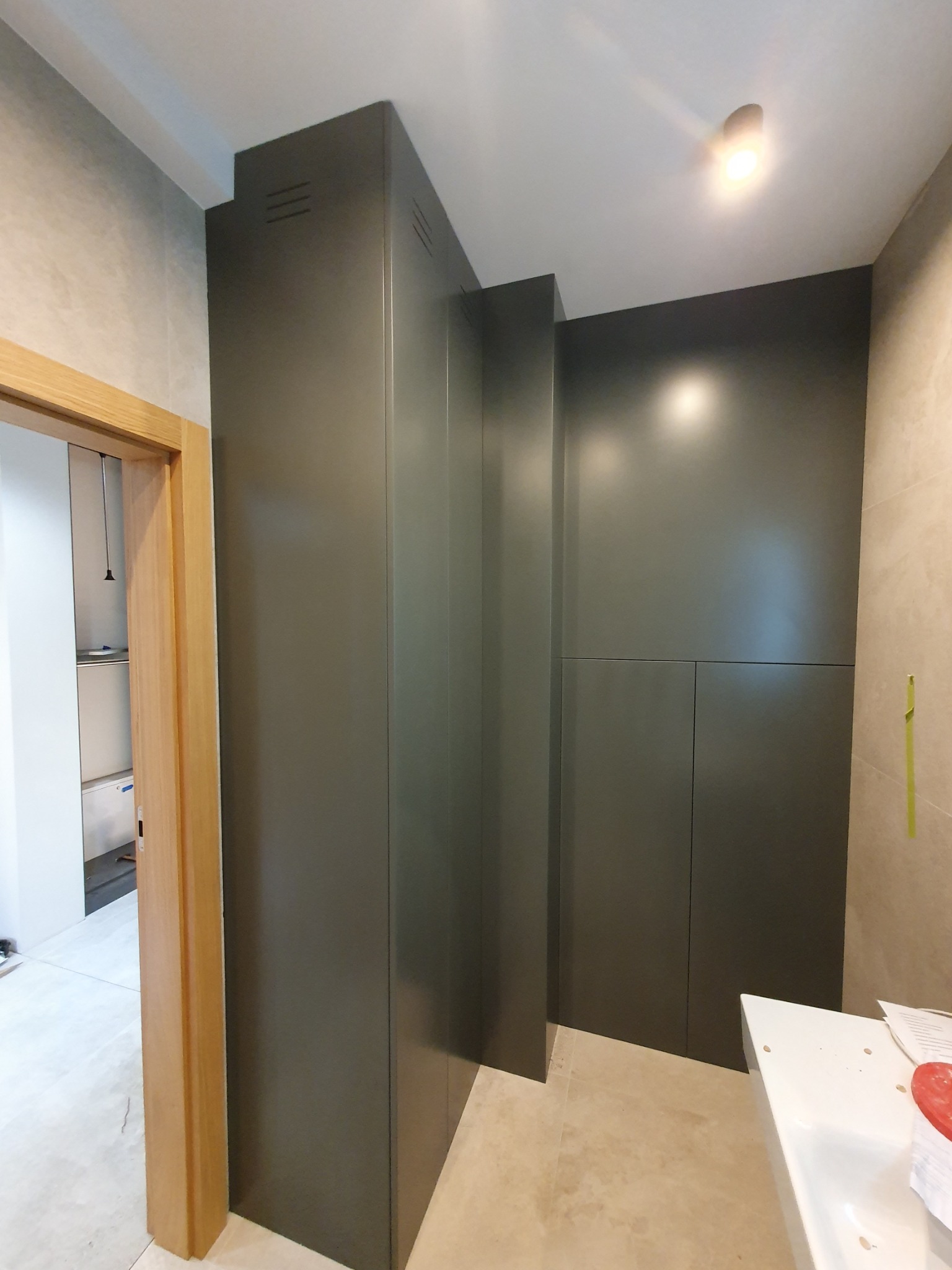 Wysoka, grafitowa szafa wnękowa z ukrytymi drzwiczkami, wkomponowana w przestrzeń łazienki z beżowymi płytkami i oświetleniem punktowym.