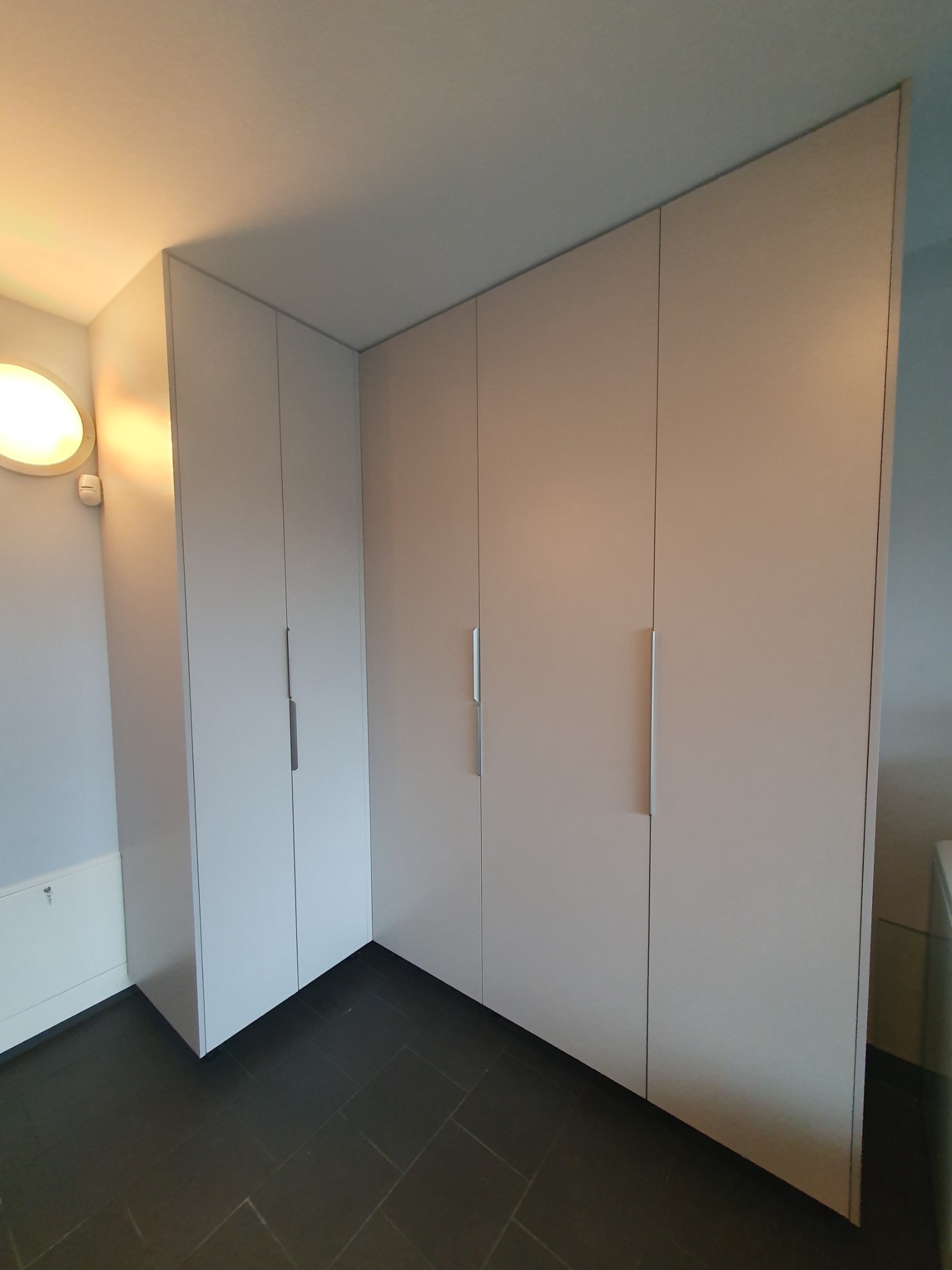 Biała szafa wnękowa z czterema drzwiami i minimalistycznymi, podłużnymi uchwytami, dopasowana do rogu pomieszczenia z ciemną podłogą.