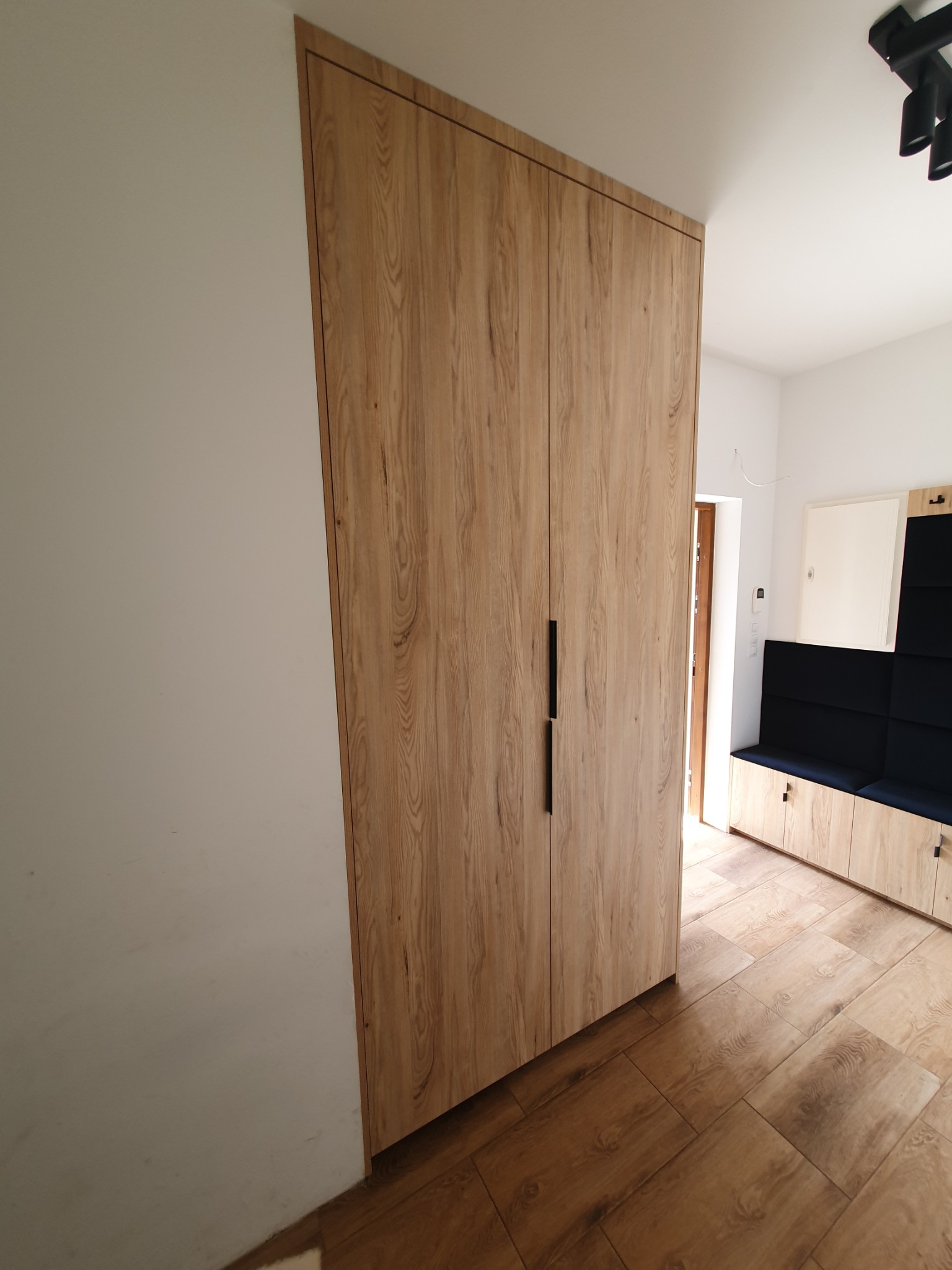 Dwudrzwiowa szafa z drewnopodobnymi frontami wkomponowana we wnękę, z czarnymi, minimalistycznymi uchwytami, widoczna z boku, z fragmentem siedziska z ciemnoniebieską tapicerką w tle.