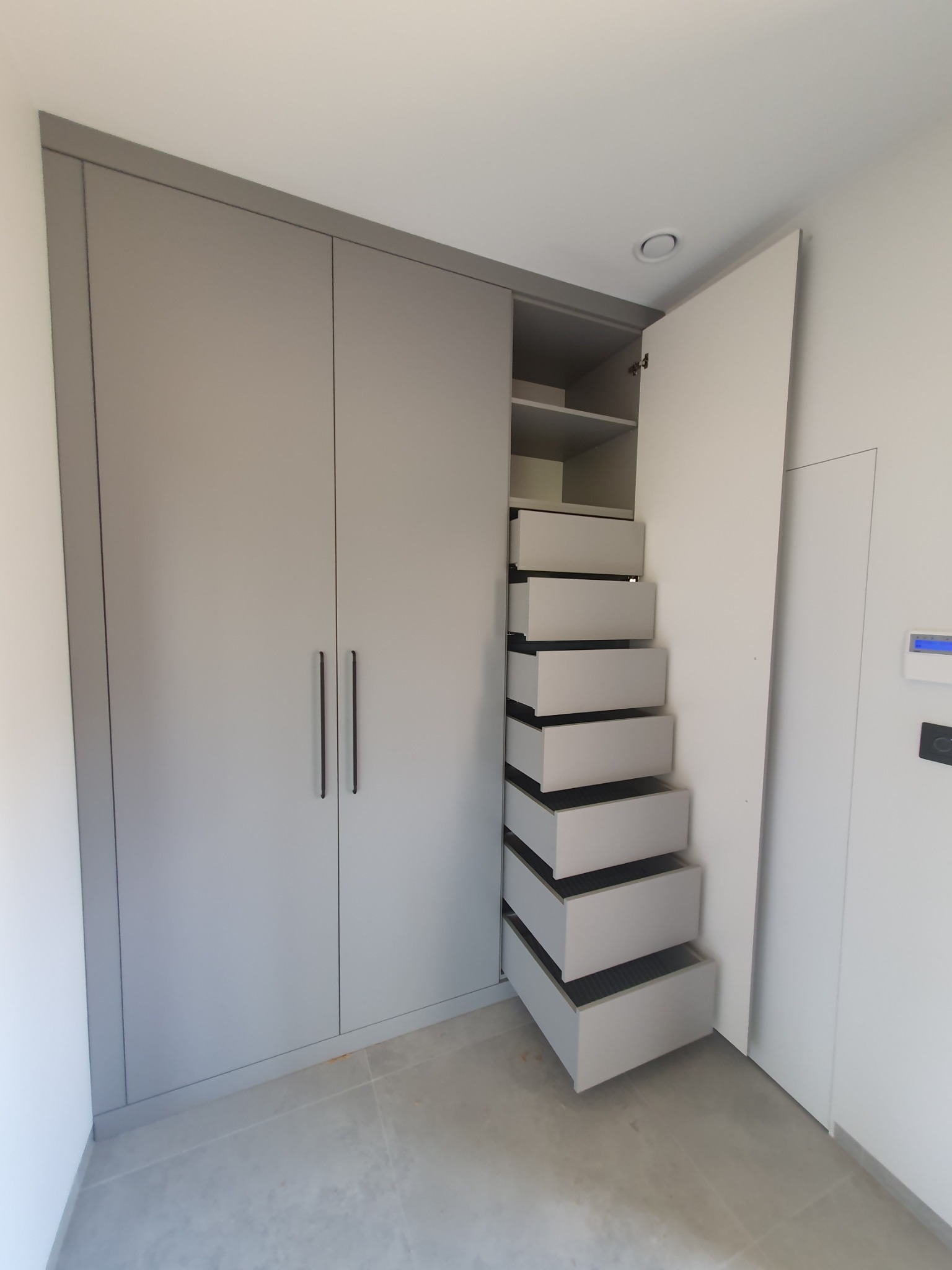 Szara szafa wnękowa z otwartymi drzwiami i szeregiem wysuwanych szuflad, widok z przodu na jasnym tle ściany i podłogi.
