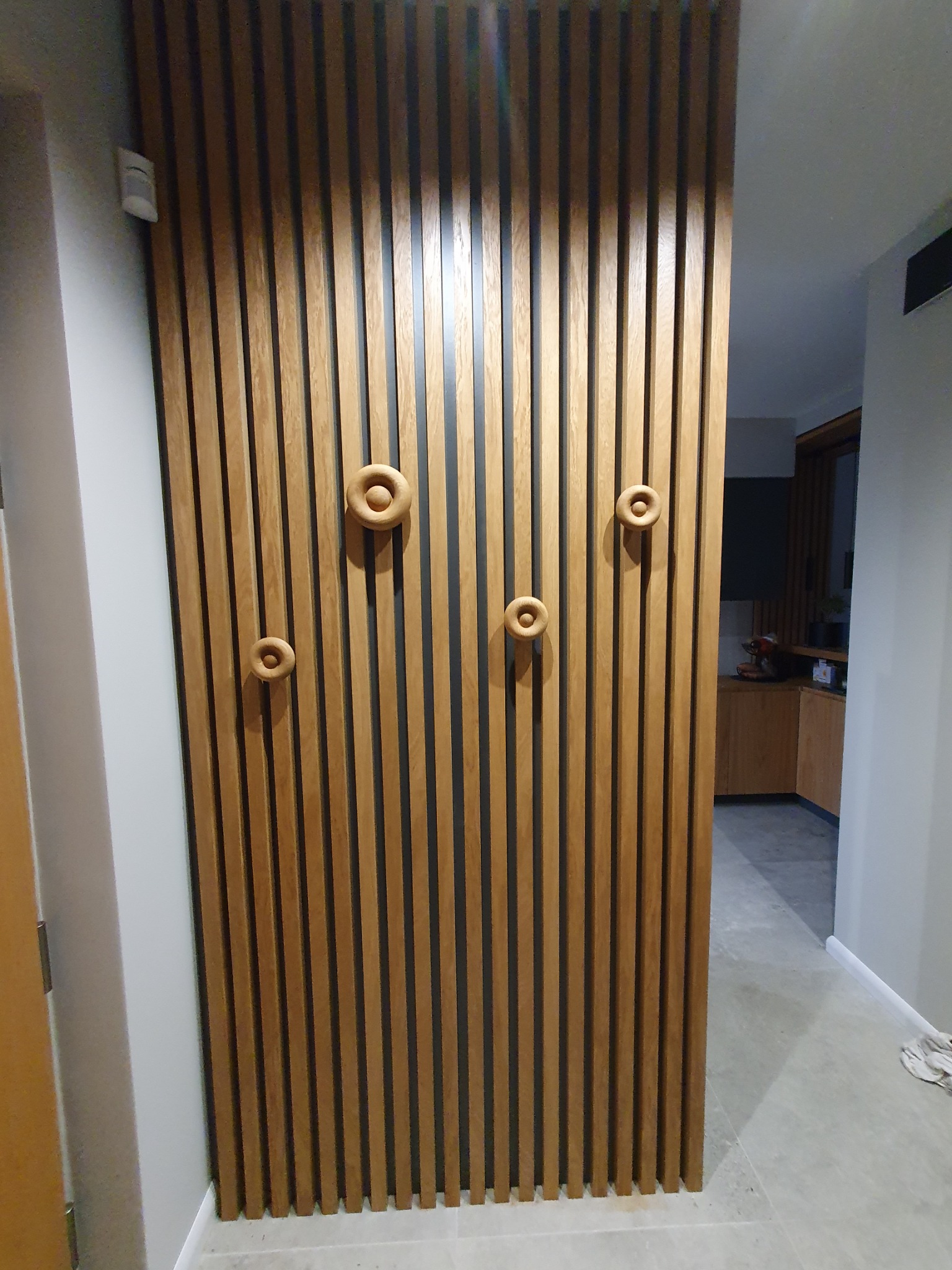 Drewniana ścianka działowa z pionowych listew w kolorze jasnego dębu, ozdobiona okrągłymi uchwytami, oddzielająca przestrzeń w nowoczesnym wnętrzu.