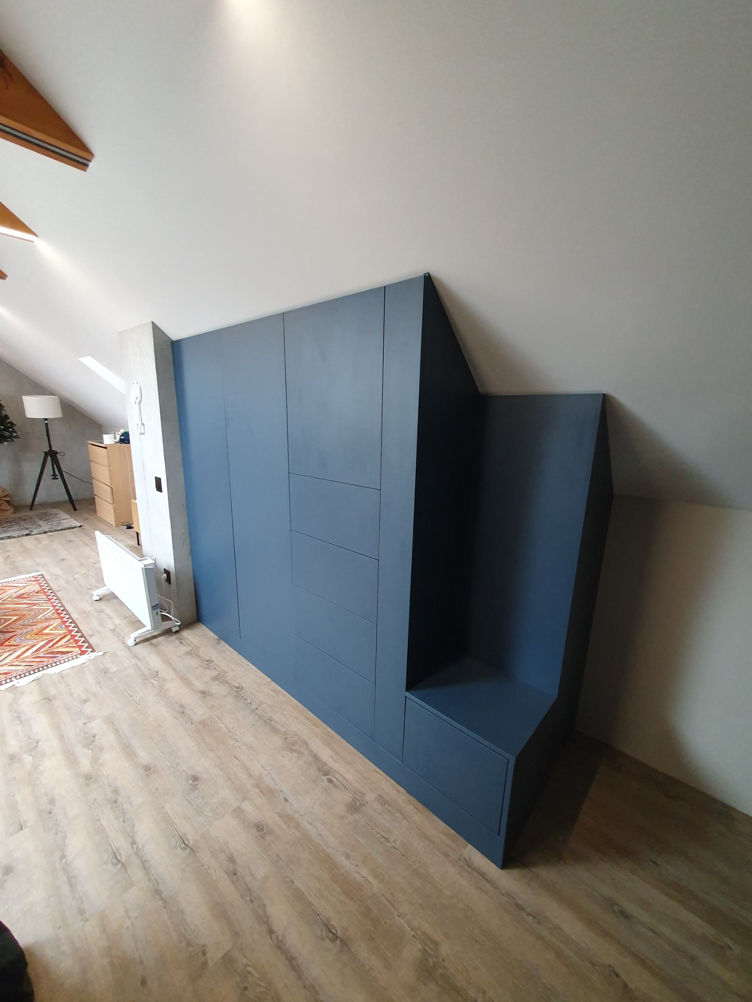 Niebieska szafa wnękowa na poddaszu ze zintegrowanym siedziskiem, widoczny fragment grzejnika i dywan w tle.