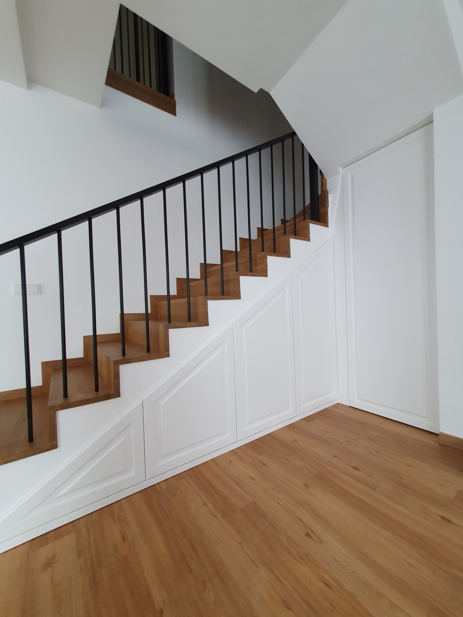 Schody z drewnianymi stopniami i czarną balustradą, zabudowane białymi szafkami z panelami dekoracyjnymi, widok z dołu na drewnianą podłogę.