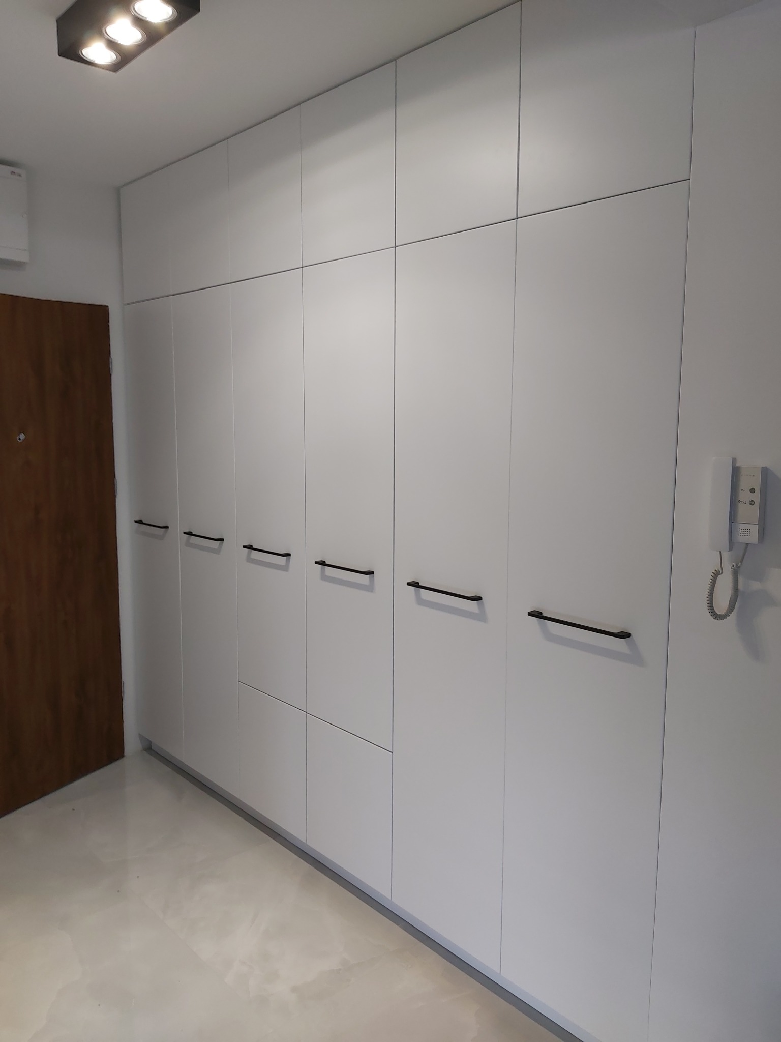 Biała, minimalistyczna szafa wnękowa z czarnymi, podłużnymi uchwytami, widoczna w przedpokoju z brązowymi drzwiami i białym domofonem na ścianie.