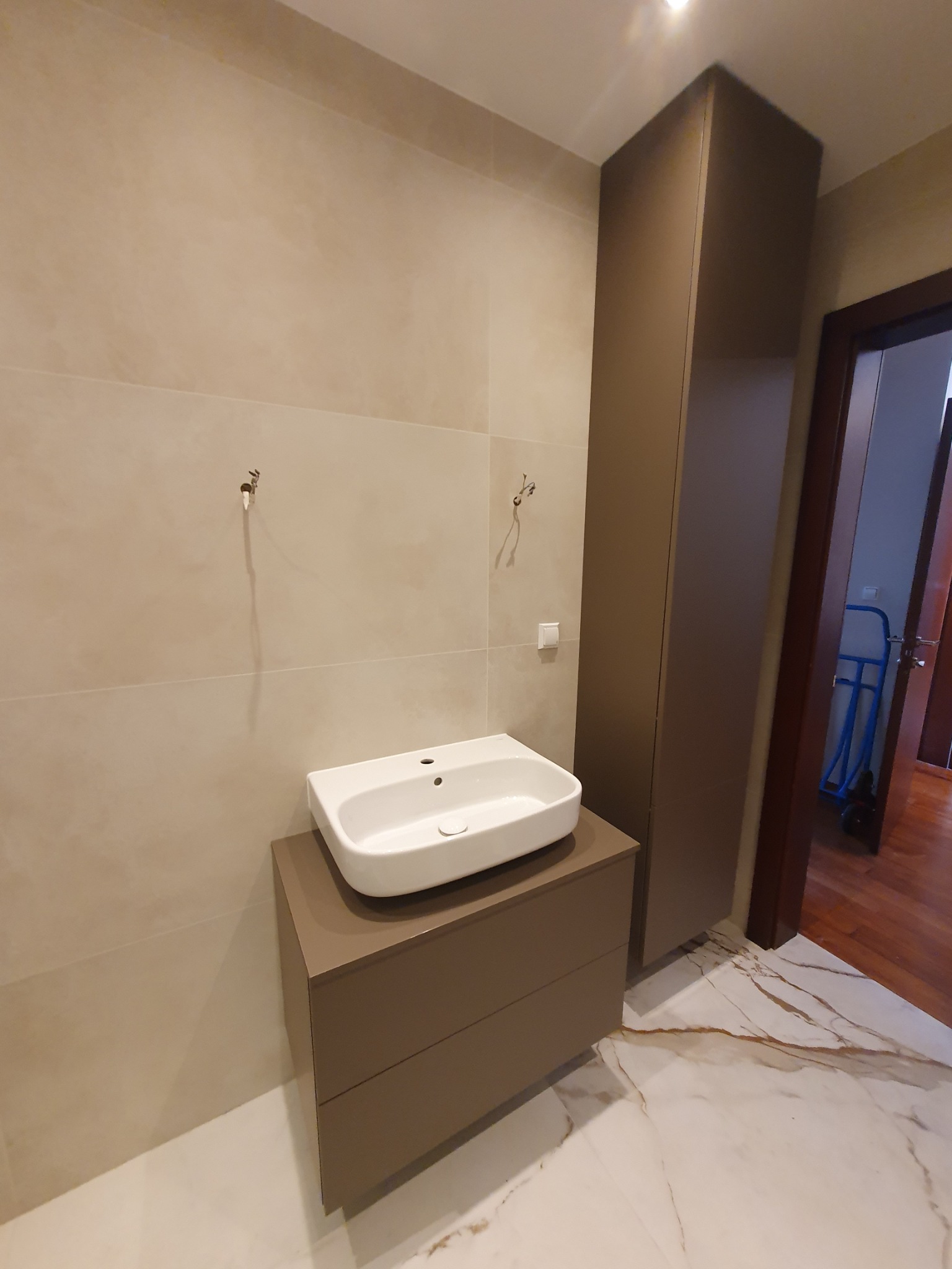 Nowoczesna łazienka z białą umywalką nablatową na szafce w kolorze taupe, wysoka szafa w tym samym kolorze, beżowe płytki ścienne i marmurowa podłoga z brązowymi żyłami, widoczny fragment otwartych...