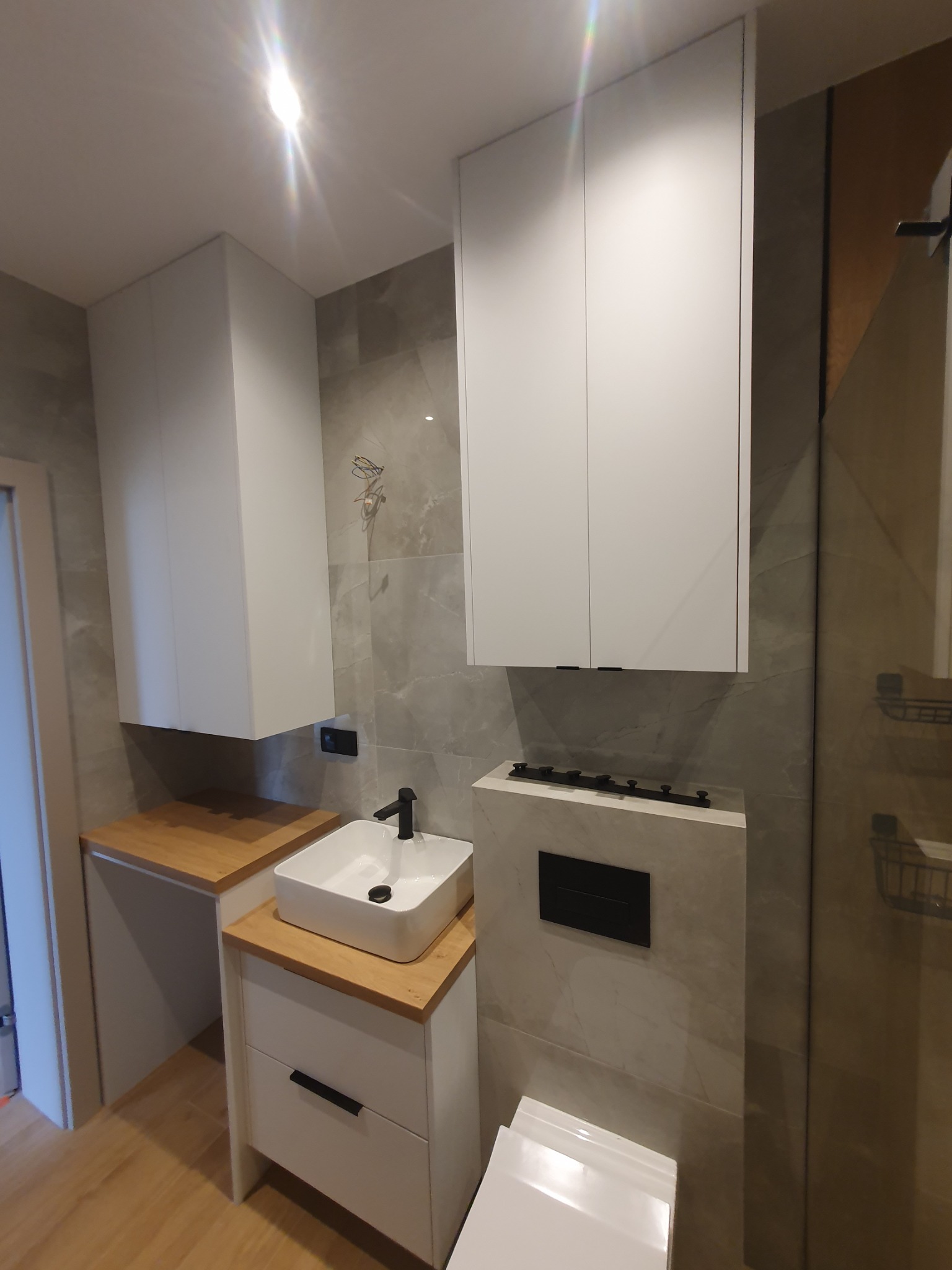 Nowoczesna łazienka z białymi szafkami wiszącymi, umywalką nablatową na blacie z jasnego drewna i szarą płytką imitującą marmur na ścianach.