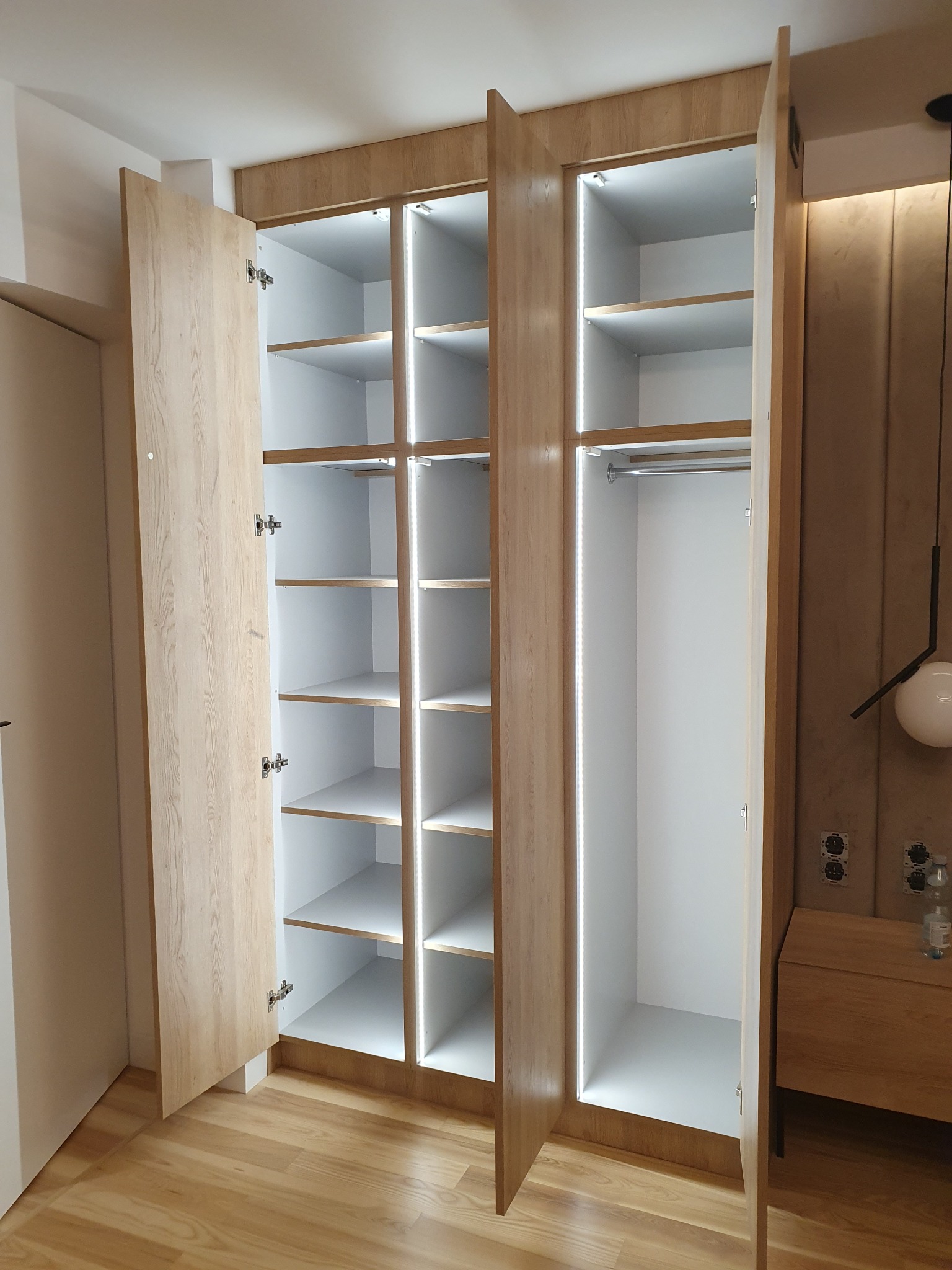 Otwarta, trzydrzwiowa szafa wnękowa z oświetleniem LED, jasne drewno i białe wnętrze, widoczne półki i drążek na ubrania, minimalistyczny design, na podłodze panele.