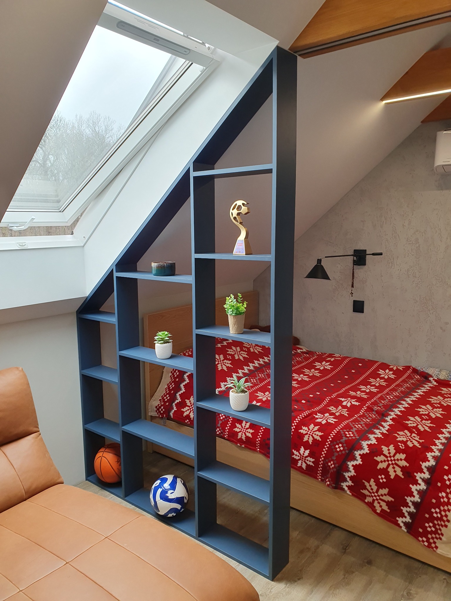 Niebieski regał z półkami w pokoju na poddaszu, oddzielający strefę wypoczynkową z skórzanym leżanką od sypialni z łóżkiem przykrytym czerwoną narzutą w śnieżynki, na półkach dekoracje i piłki.