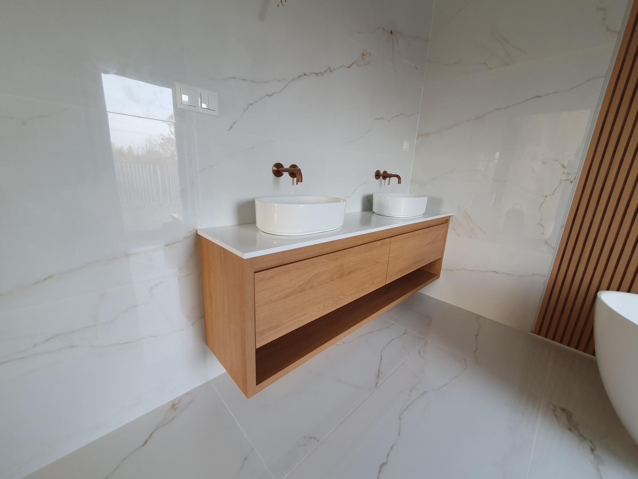 Nowoczesna łazienka z drewnianą szafką pod umywalki nablatowe, miedziane baterie, białe marmurowe płytki na ścianach i podłodze, oraz dekoracyjny panel z drewnianych listew.