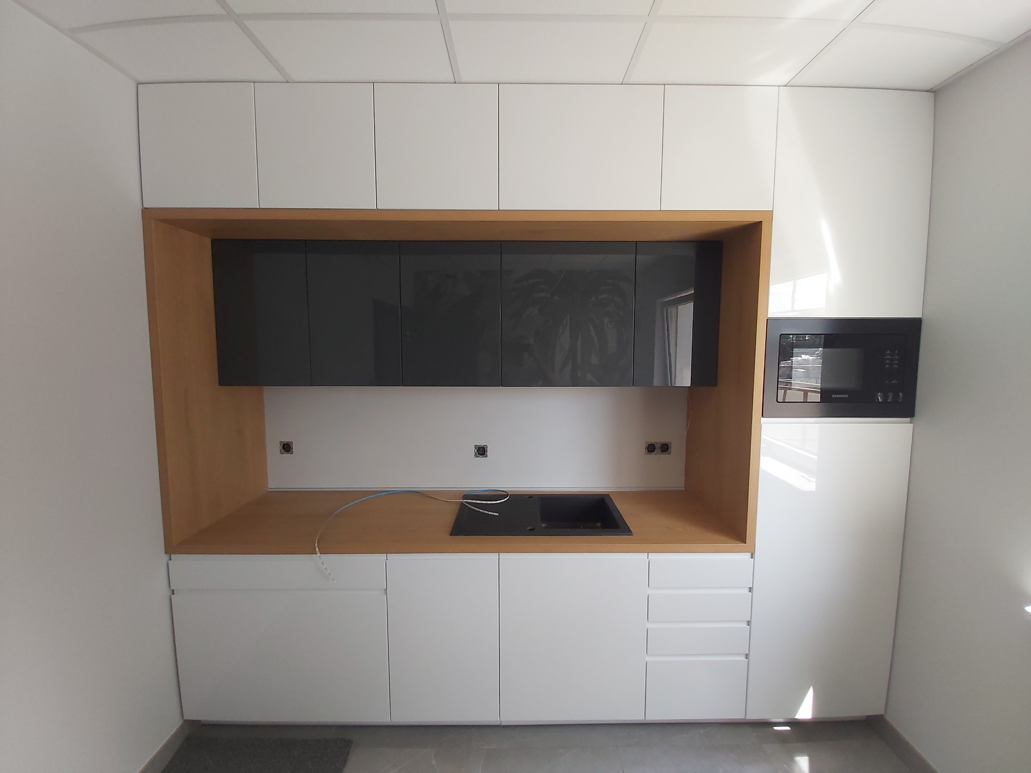 Nowoczesna zabudowa kuchenna z białymi szafkami górnymi i dolnymi, drewnianą wnęką z szarymi szafkami i czarnym zlewem, oraz mikrofalówką po prawej stronie, widoczne kable i gniazdka.