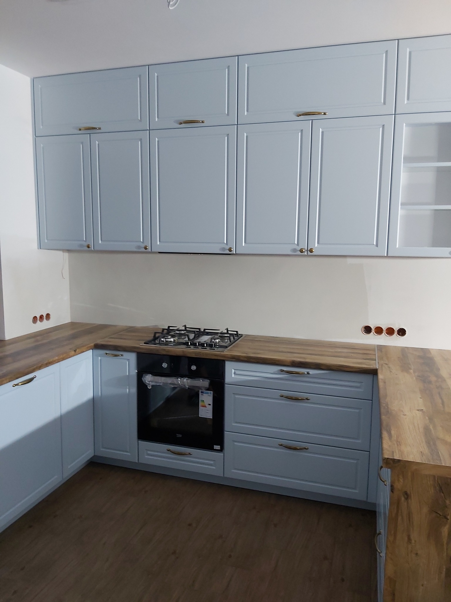 Jasnoniebieska zabudowa kuchenna z drewnianym blatem, widoczne górne szafki z przeszkloną witryną, płyta gazowa i piekarnik, minimalistyczne złote uchwyty.