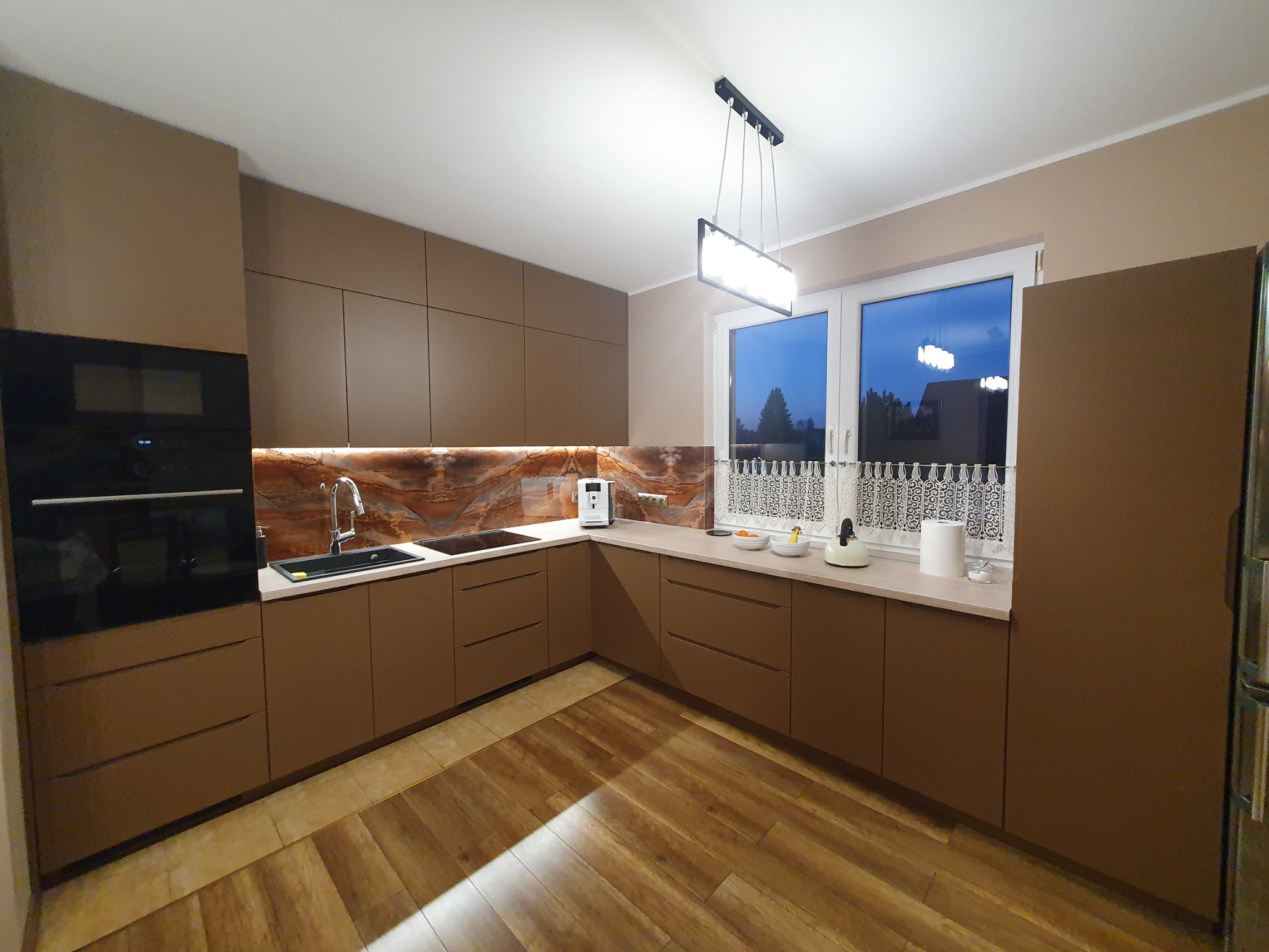 Nowoczesna kuchnia w odcieniach brązu z marmurkowym backsplashiem, minimalistycznym oświetleniem i drewnianą podłogą.