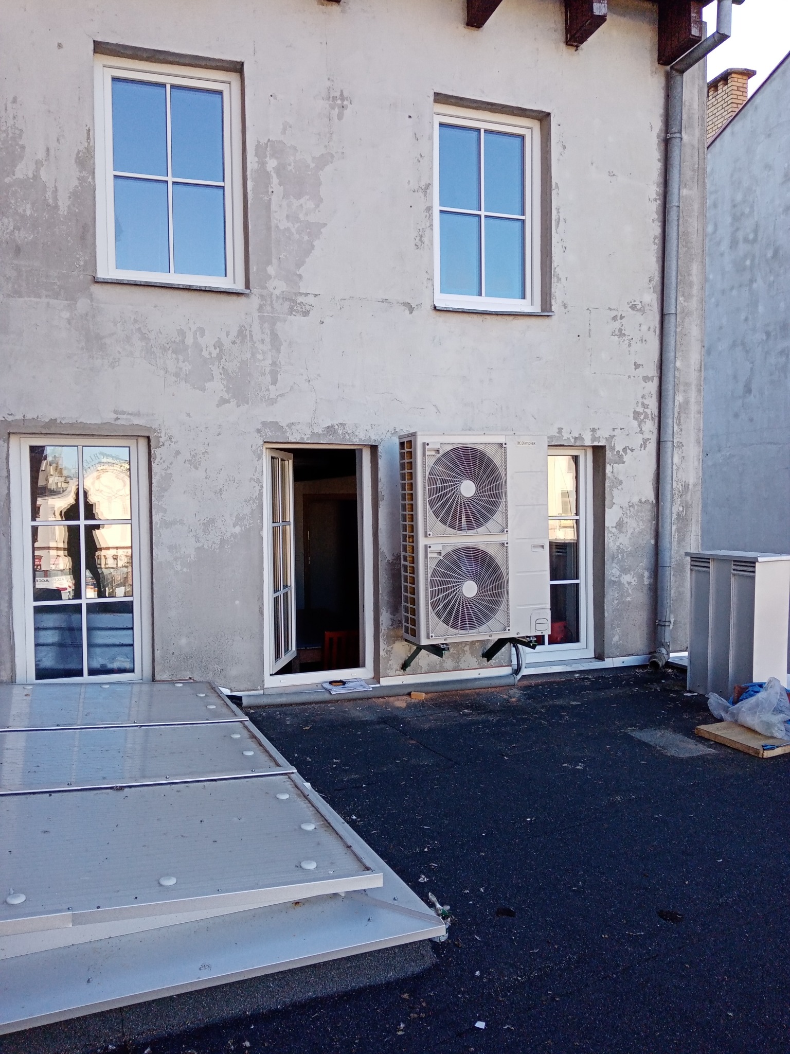 Zewnętrzna jednostka pompy ciepła Dimplex typu split zamontowana na elewacji budynku, widoczna obok otwartego okna, z fragmentem dachu i instalacją odprowadzającą wodę.