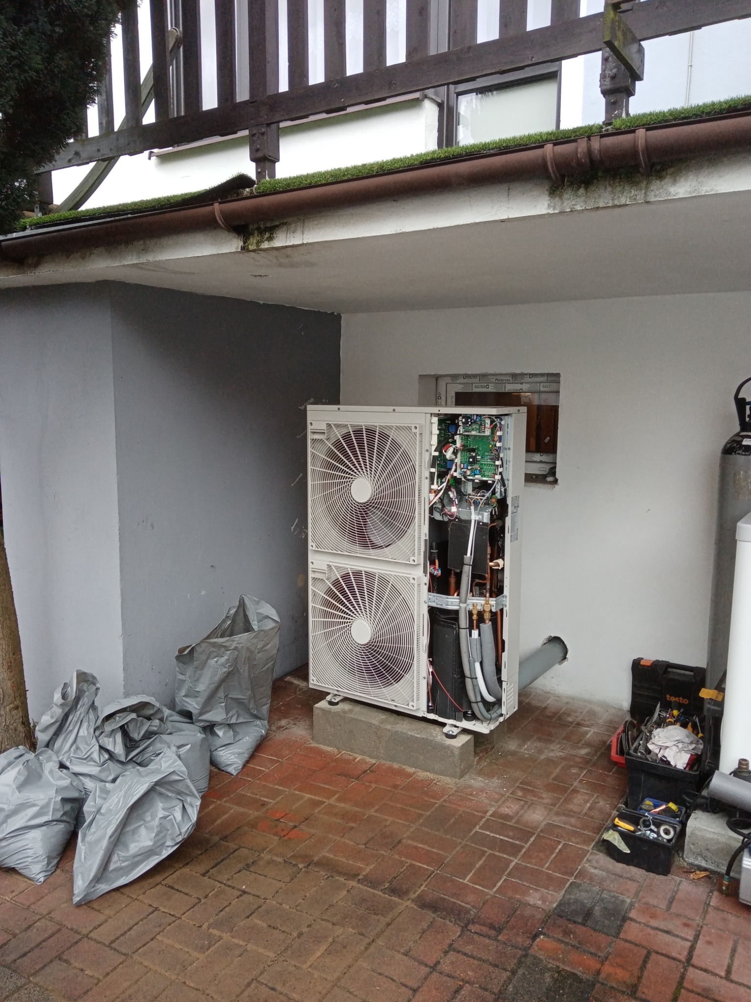 Otwarta dwuwentylatorowa jednostka zewnętrzna z widocznymi komponentami wewnętrznymi, stojąca na betonowym fundamencie na ceglanym patio, obok narzędzi i worków na śmieci.