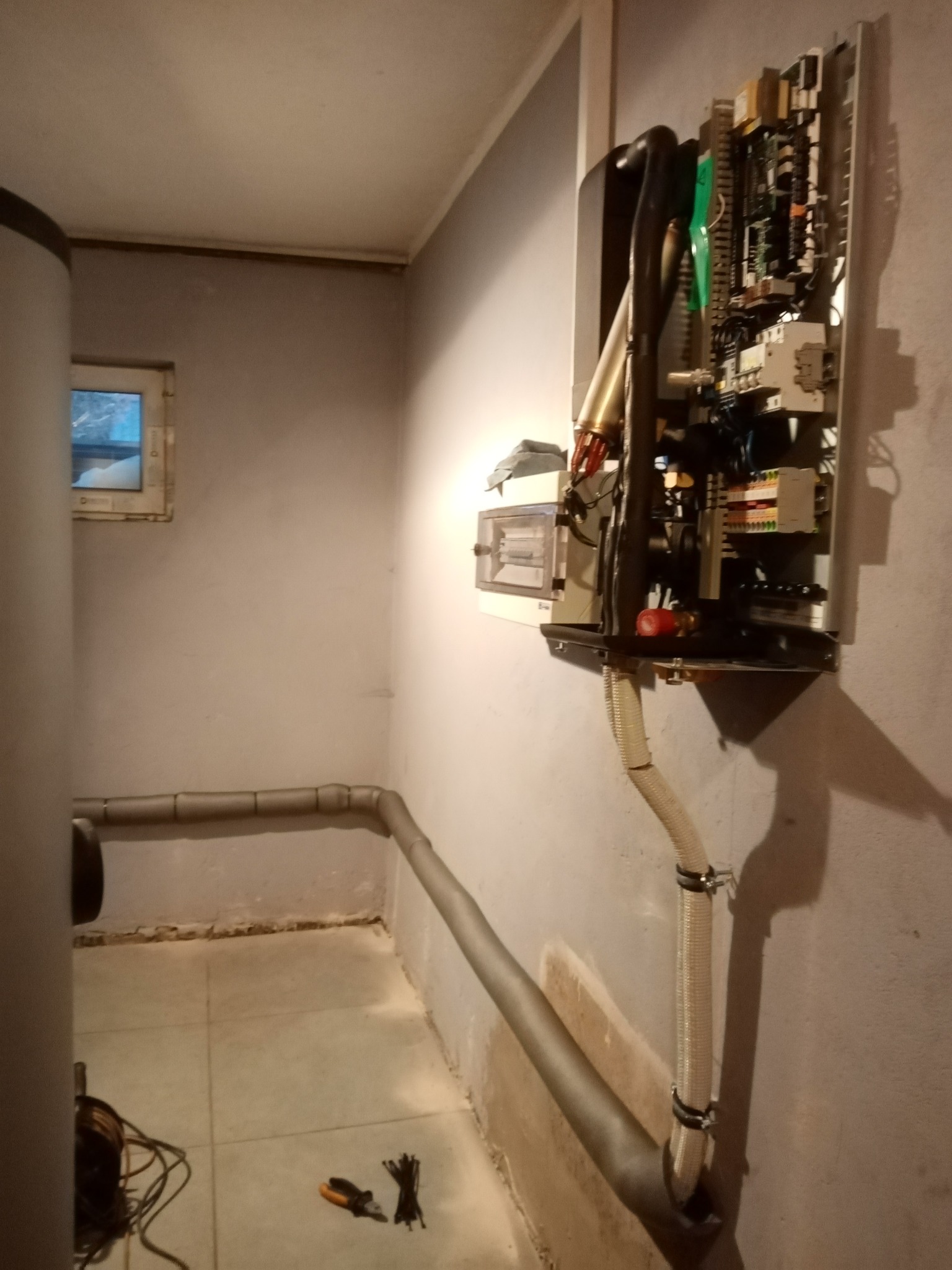 Otwarta obudowa urządzenia grzewczego na ścianie w piwnicy, widoczne wewnętrzne komponenty elektroniczne i hydrauliczne, rury instalacyjne z izolacją termiczną, narzędzia i opaski zaciskowe...