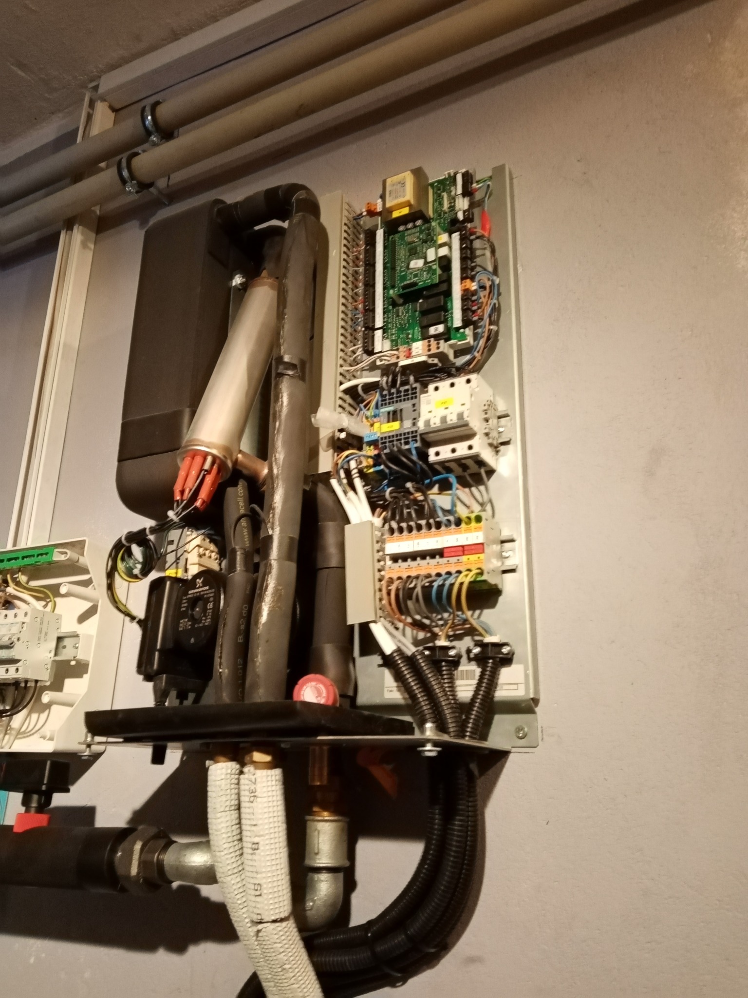 Otwarta obudowa pompy ciepła z widocznymi elementami elektronicznymi, przewodami i rurami, zainstalowana przy szarej ścianie.