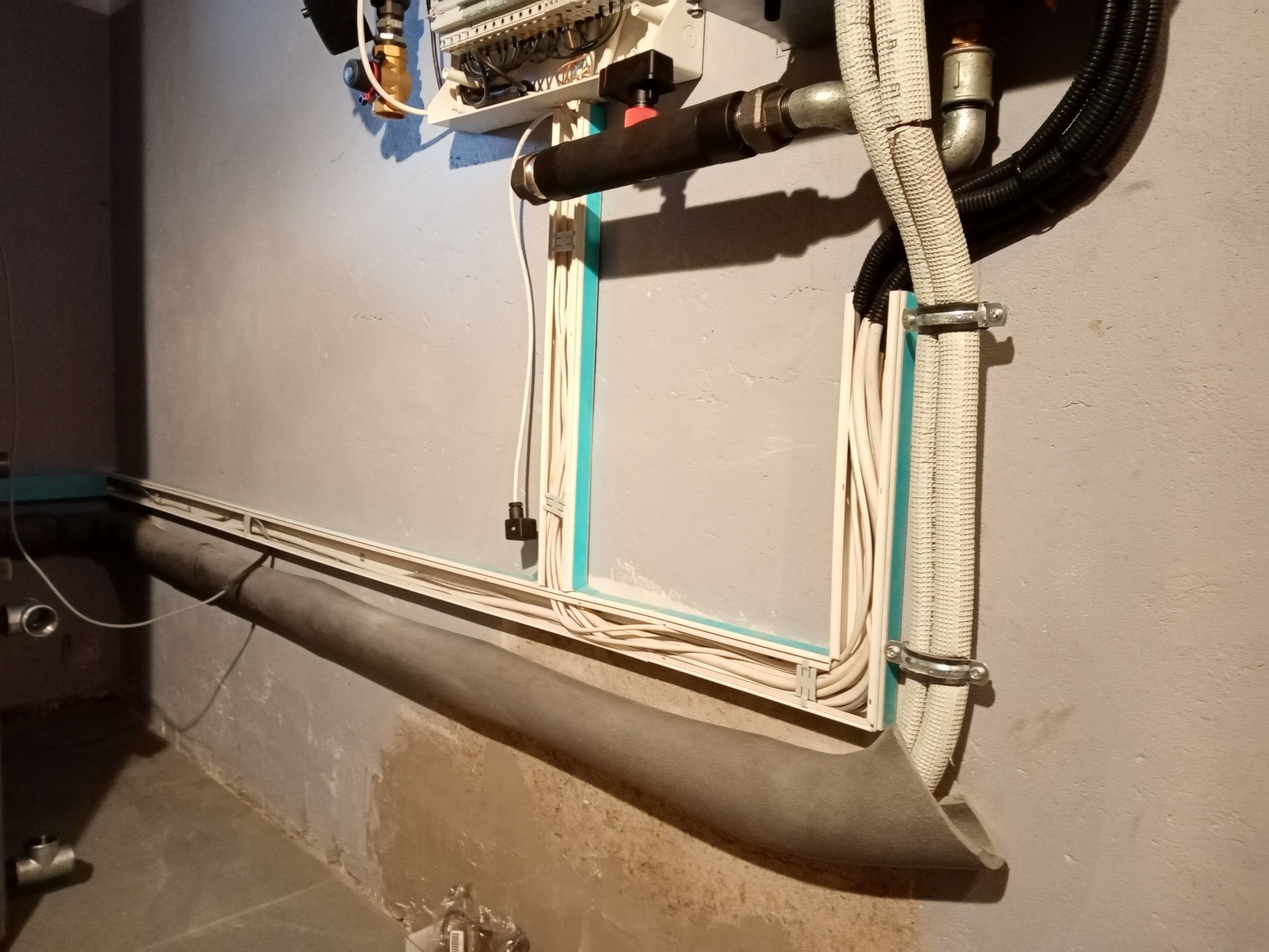 Szare pomieszczenie techniczne z instalacją: rury w izolacji, przewody elektryczne w listwach, rozdzielnica elektryczna na ścianie.