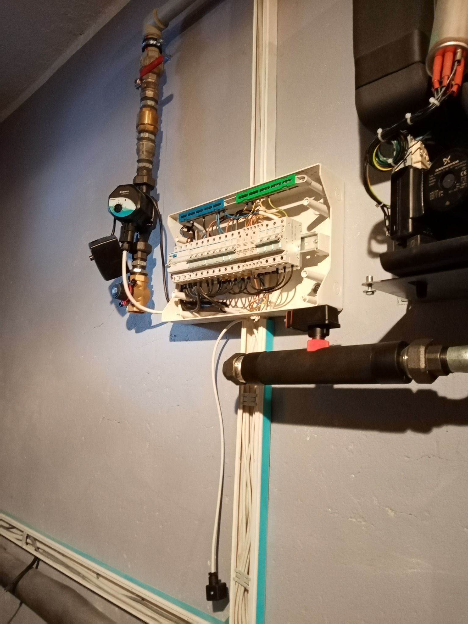Szara ściana z zamontowaną skrzynką elektryczną z widocznymi przewodami, rurami instalacyjnymi i licznikiem ciepła z zaworem regulacyjnym.