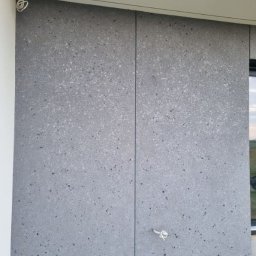 STAFF-BUD - Szara elewacja budynku z widocznymi łączeniami płyt i wystającymi przewodami elektrycznymi. Fragment okna po prawej stronie. Widoczne elementy konstrukcyjne u dołu.