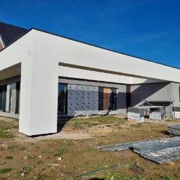 STAFF-BUD - Nowoczesny dom jednorodzinny w trakcie prac elewacyjnych. Widoczne rusztowania i materiały budowlane. Elewacja w kolorze białym, okna zabezpieczone folią.