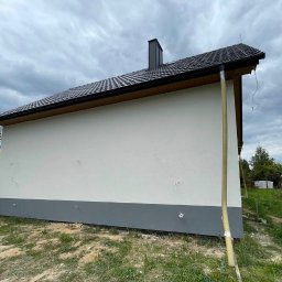 STAFF-BUD - Świeżo otynkowana ściana domu z widocznymi rurami spustowymi i fragmentem dachu z kominem na tle pochmurnego nieba i zieleni.