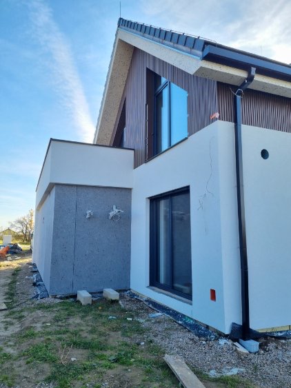 Nowoczesny dom z elewacją częściowo ocieploną szarym styropianem, górna część wykończona drewnianą okładziną. Widoczne okno i rynna. Prace w toku.