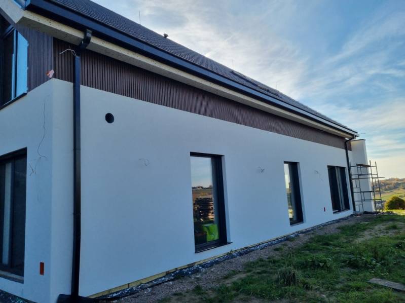 Biała elewacja domu z brązowymi panelami, czarnymi ramami okiennymi i rynnami. Widoczne rusztowanie i trawa. Świeżo położony tynk, widoczne ślady po łataniu.