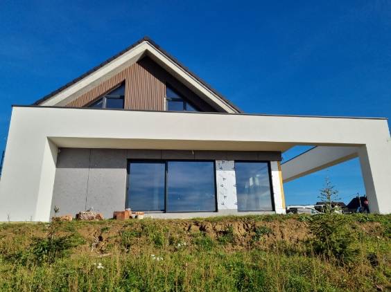 Nowoczesny dom z częściowo ocieploną elewacją. Widoczne szare płyty izolacyjne i białe wykończenie. Duże okna odbijają błękit nieba. Ujęcie z poziomu gruntu.