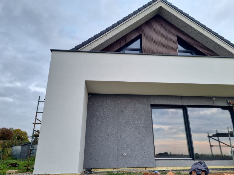 Nowoczesna elewacja domu z białym tynkiem i szarą okładziną. Widoczne okno z odbiciem nieba i drabina oparta o ścianę. Elementy wykończeniowe w kolorze antracytowym.