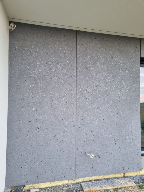 Szara elewacja budynku z widocznymi łączeniami płyt i wystającymi przewodami elektrycznymi. Fragment okna po prawej stronie. Widoczne elementy konstrukcyjne u dołu.