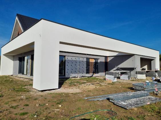 Nowoczesny dom jednorodzinny w trakcie prac elewacyjnych. Widoczne rusztowania i materiały budowlane. Elewacja w kolorze białym, okna zabezpieczone folią.