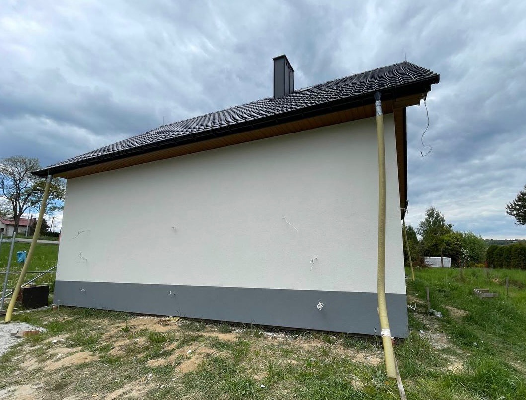 Świeżo otynkowana ściana domu z widocznymi rurami spustowymi i fragmentem dachu z kominem na tle pochmurnego nieba i zieleni.