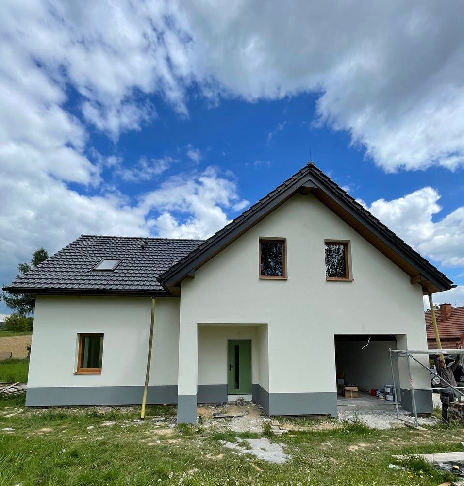 Biały dom z szarą podmurówką i zielonymi drzwiami garażowymi, widok z dołu na tle błękitnego nieba z chmurami. Widoczne rusztowania w garażu.