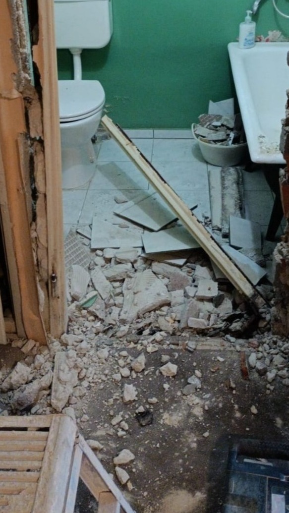 Zdemolowana łazienka w trakcie prac remontowych. Gruz, potłuczone płytki i uszkodzona futryna. Widoczne elementy wyposażenia: toaleta, umywalka. Zielona ściana w tle.