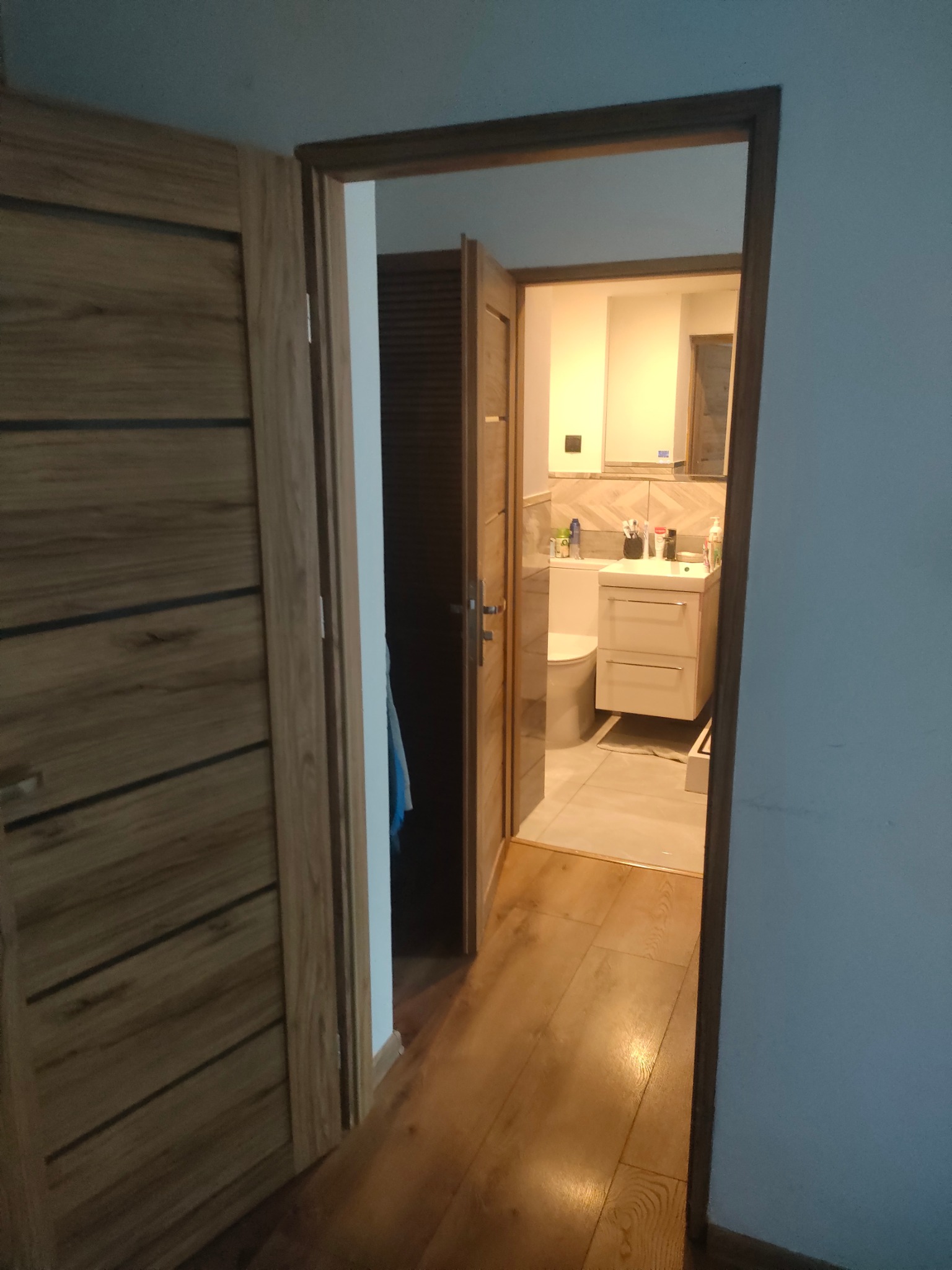 Otwarta drewniana rama drzwiowa z widokiem na wyremontowaną łazienkę z umywalką, toaletą i lustrem. Podłoga wyłożona jasnymi płytkami, ciepłe oświetlenie.