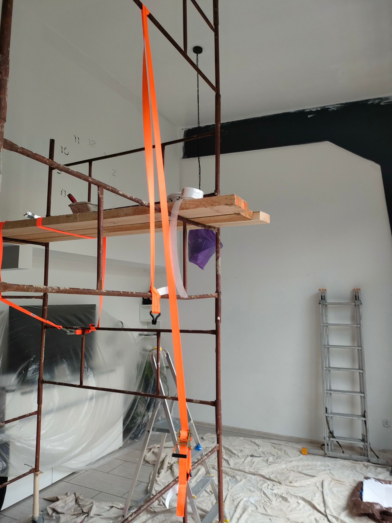 Wnętrze w trakcie prac: rusztowanie z narzędziami, zabezpieczona folią podłoga i ściana z fragmentem pomalowanej na czarno górnej części. Widoczna drabina.